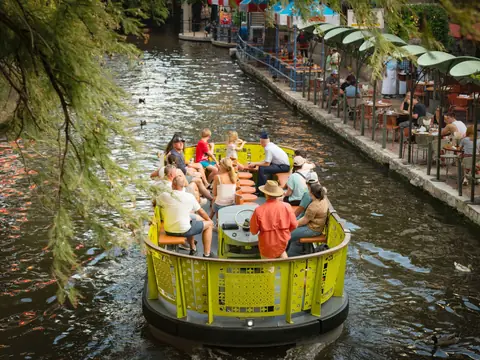 A tour boat on the green water of a river walk lined with cafes and cypress trees