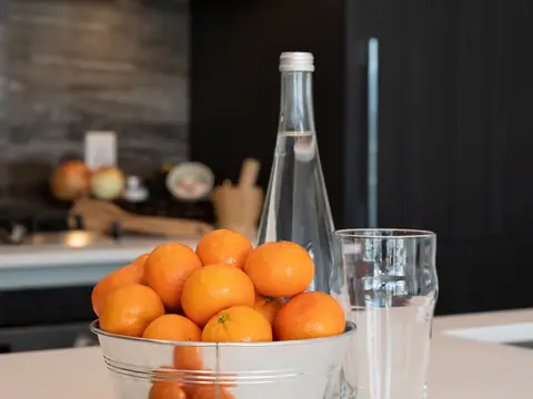 A glass and a bottle of water beside a bowl of oranges on a kitchen counter