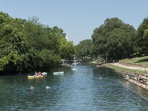 People floating on inner tubes down the Comal River past a tree-lined park in New Braunfels, Texas